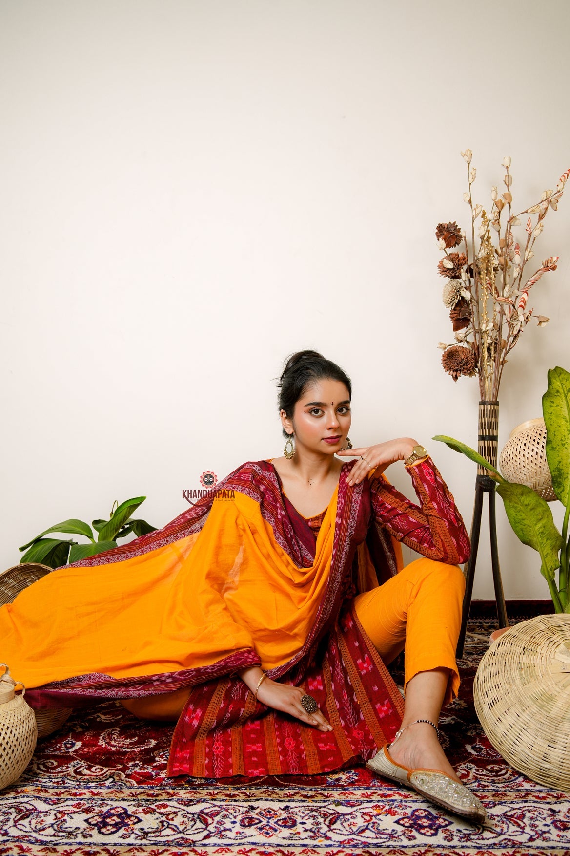 Woman in a traditional outfit sitting on a patterned rug with plants and decorative items in the background.