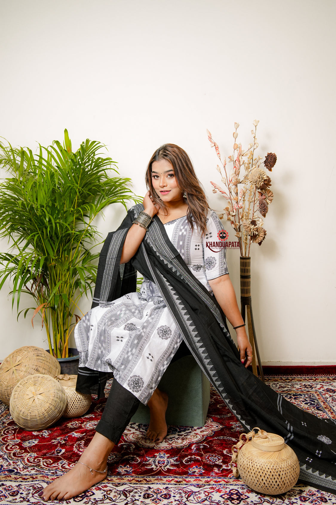 Woman sitting on a patterned rug with a long black and white scarf draped over her shoulders, surrounded by decorative items.