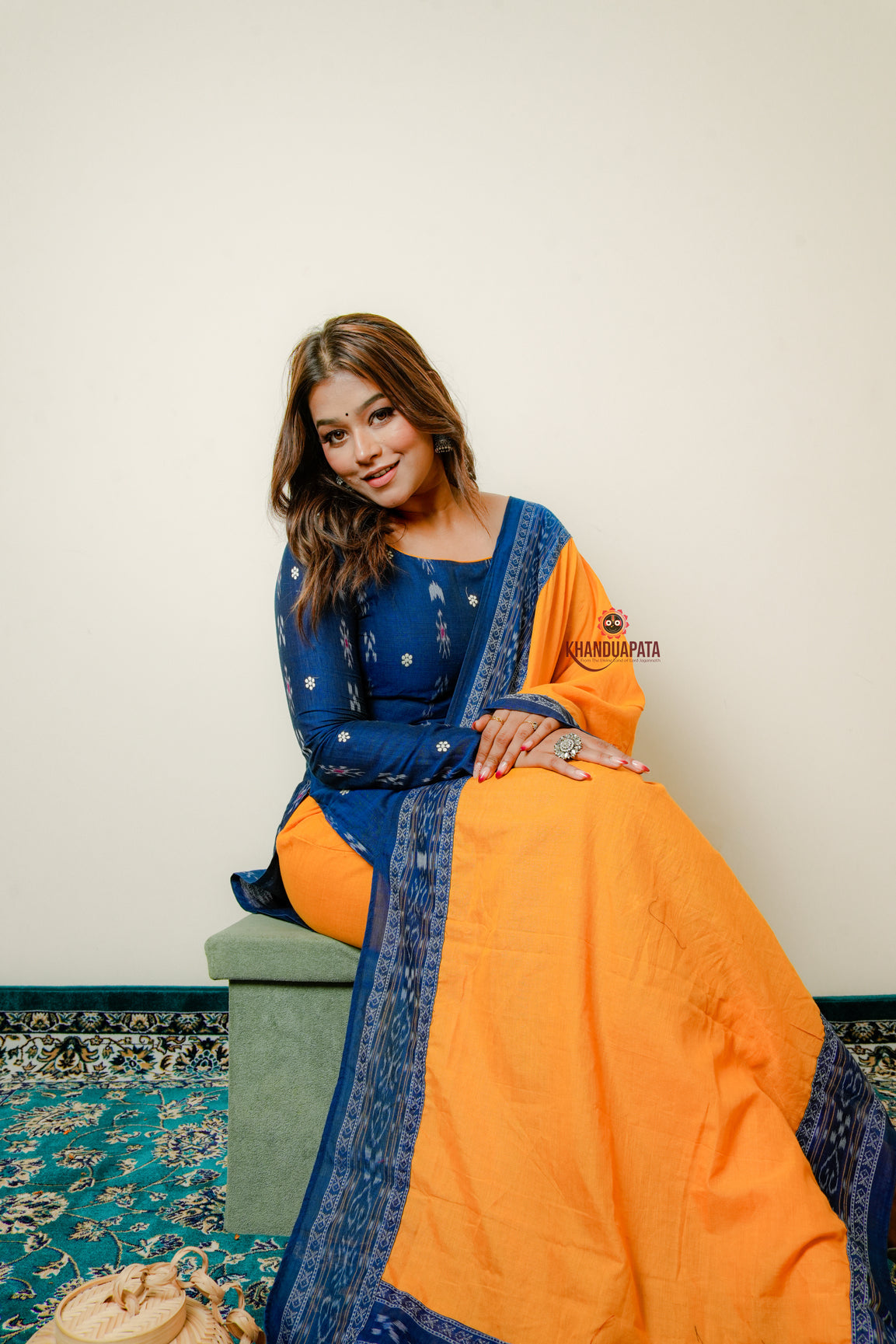 Woman wearing a blue and orange traditional outfit sitting on a patterned rug.