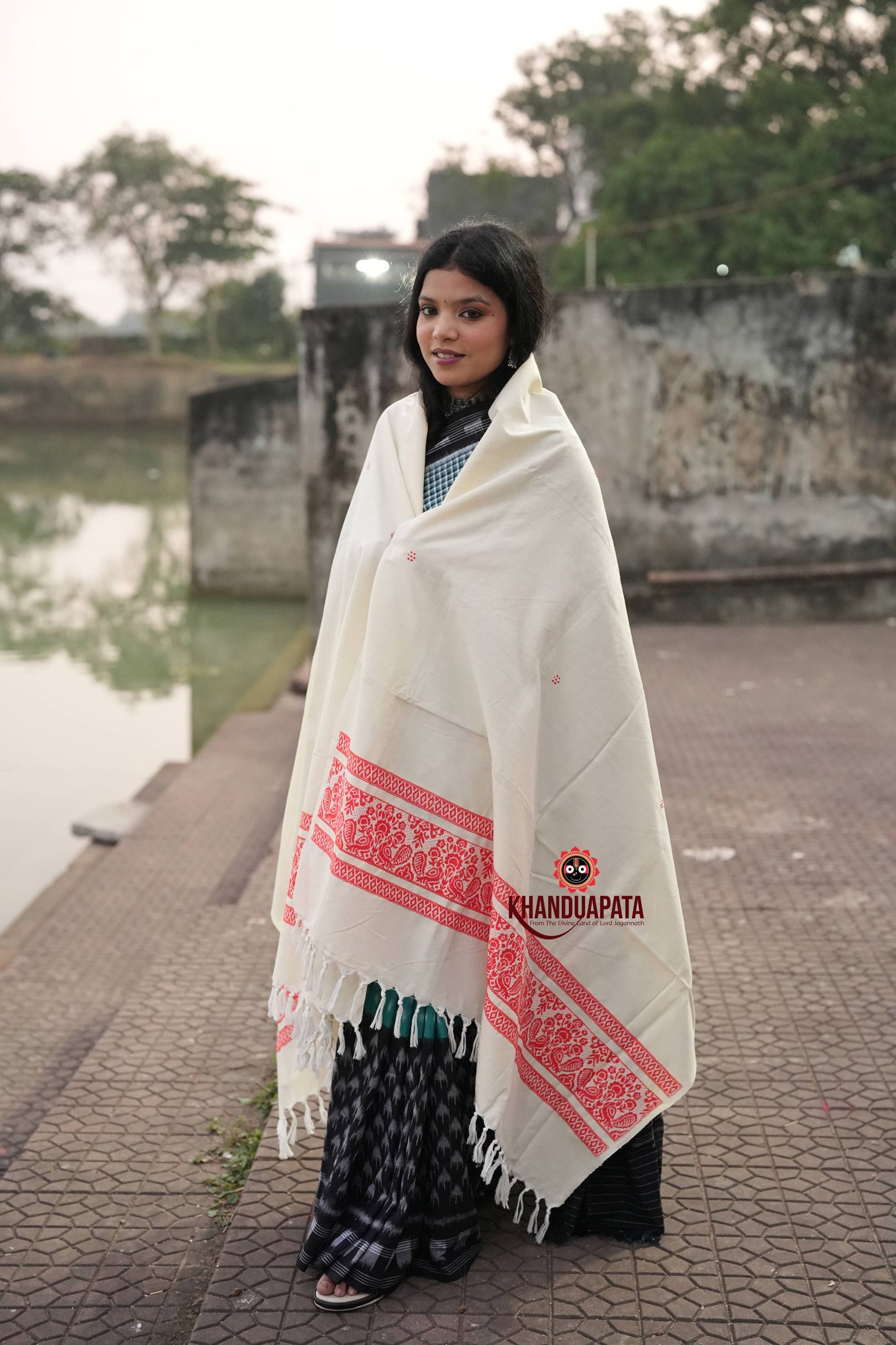 Woman wearing a white shawl with red patterns and a visible brand logo, standing by a body of water.