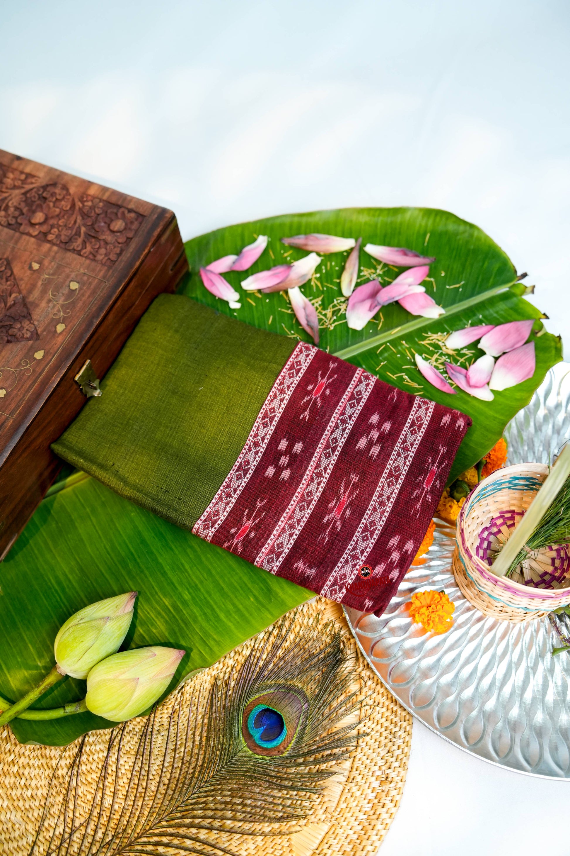 Traditional maroon saree with white patterns on a green leaf, surrounded by lotus flowers and a wooden box.