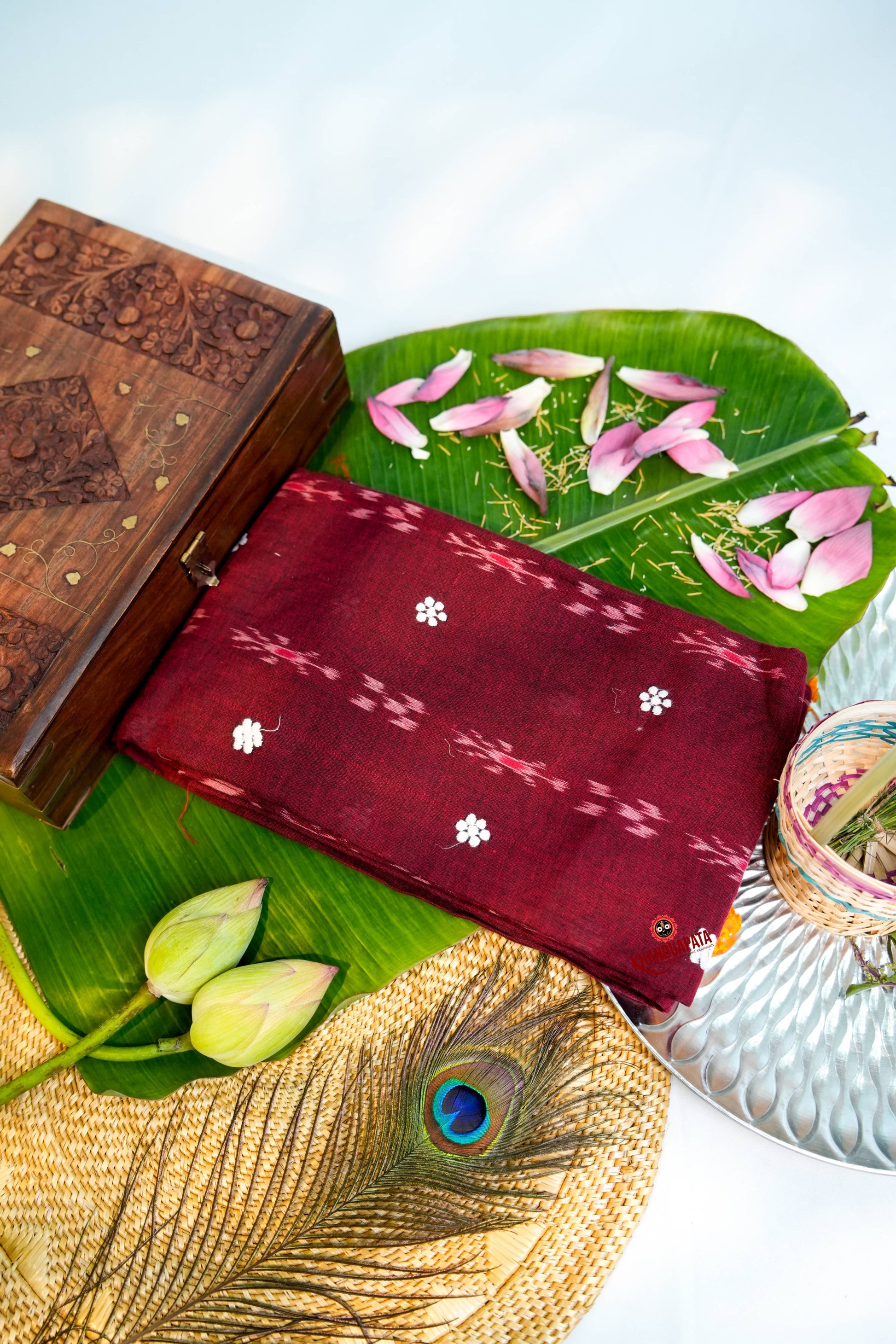Maroon fabric with white patterns on a green leaf, wooden box, and peacock feather.