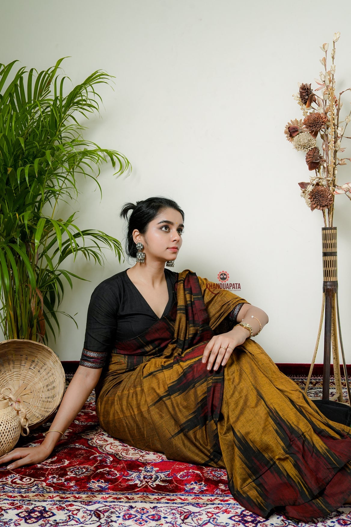 Woman in a traditional saree sitting on a patterned rug with plants and decorative items in the background.