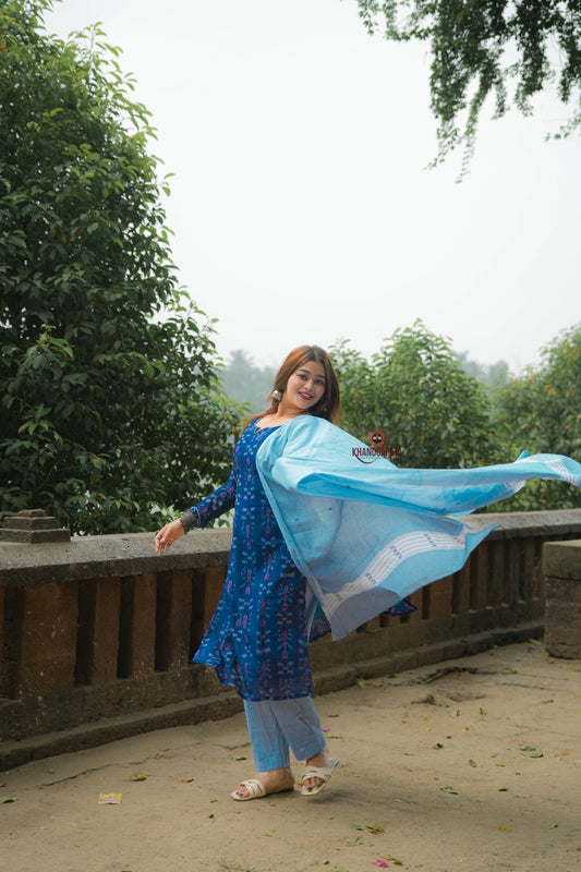 Woman in a blue traditional outfit with a flowing scarf standing outdoors with greenery in the background