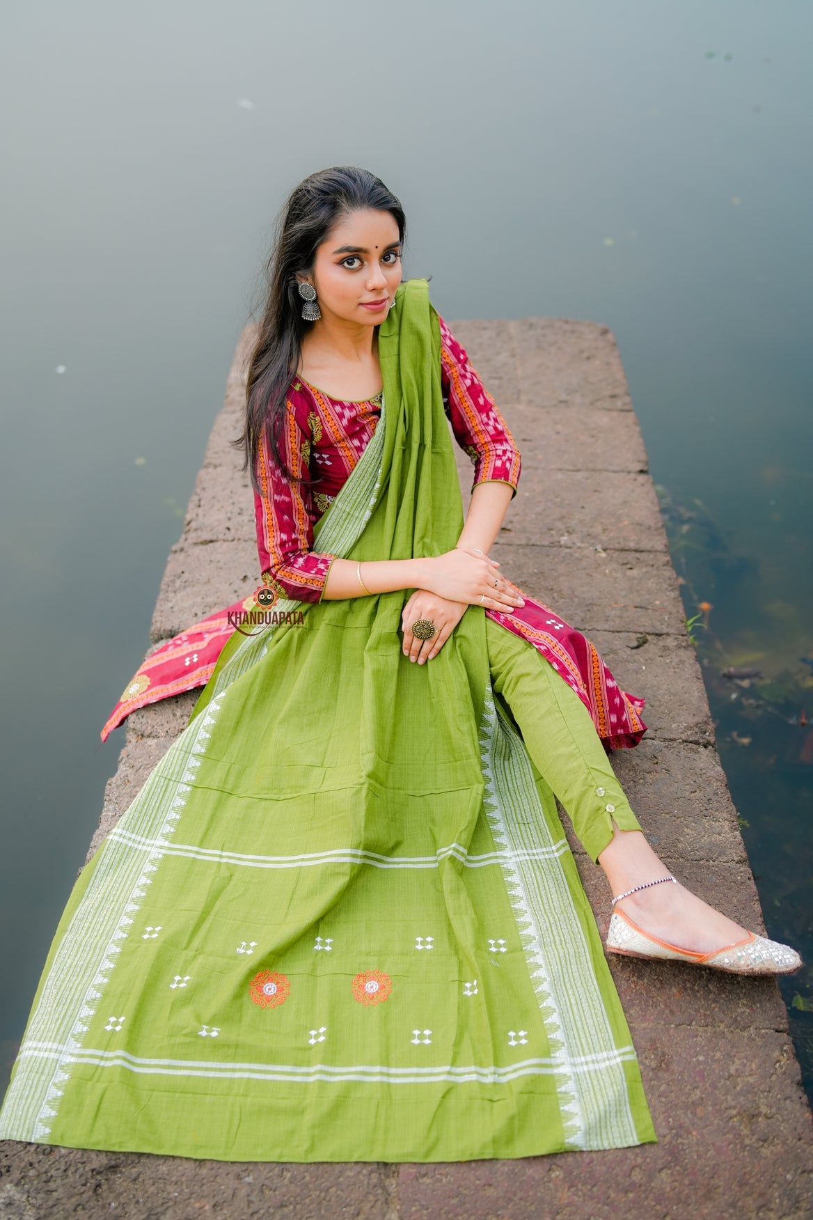 Woman in a green and pink traditional outfit sitting on a stone pier by water.