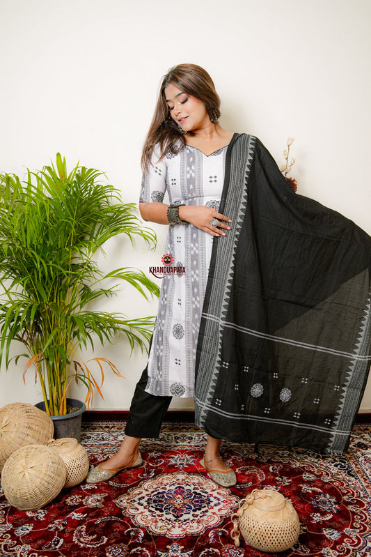 Woman holding a black saree with a white background, standing on a patterned rug with plants and decor items.
