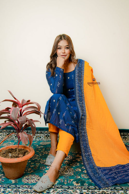 Woman in blue and orange traditional outfit sitting on a patterned rug with a plant beside her.