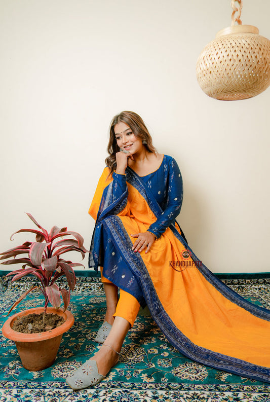 Woman in a blue and orange traditional outfit sitting on a patterned rug with a plant nearby.