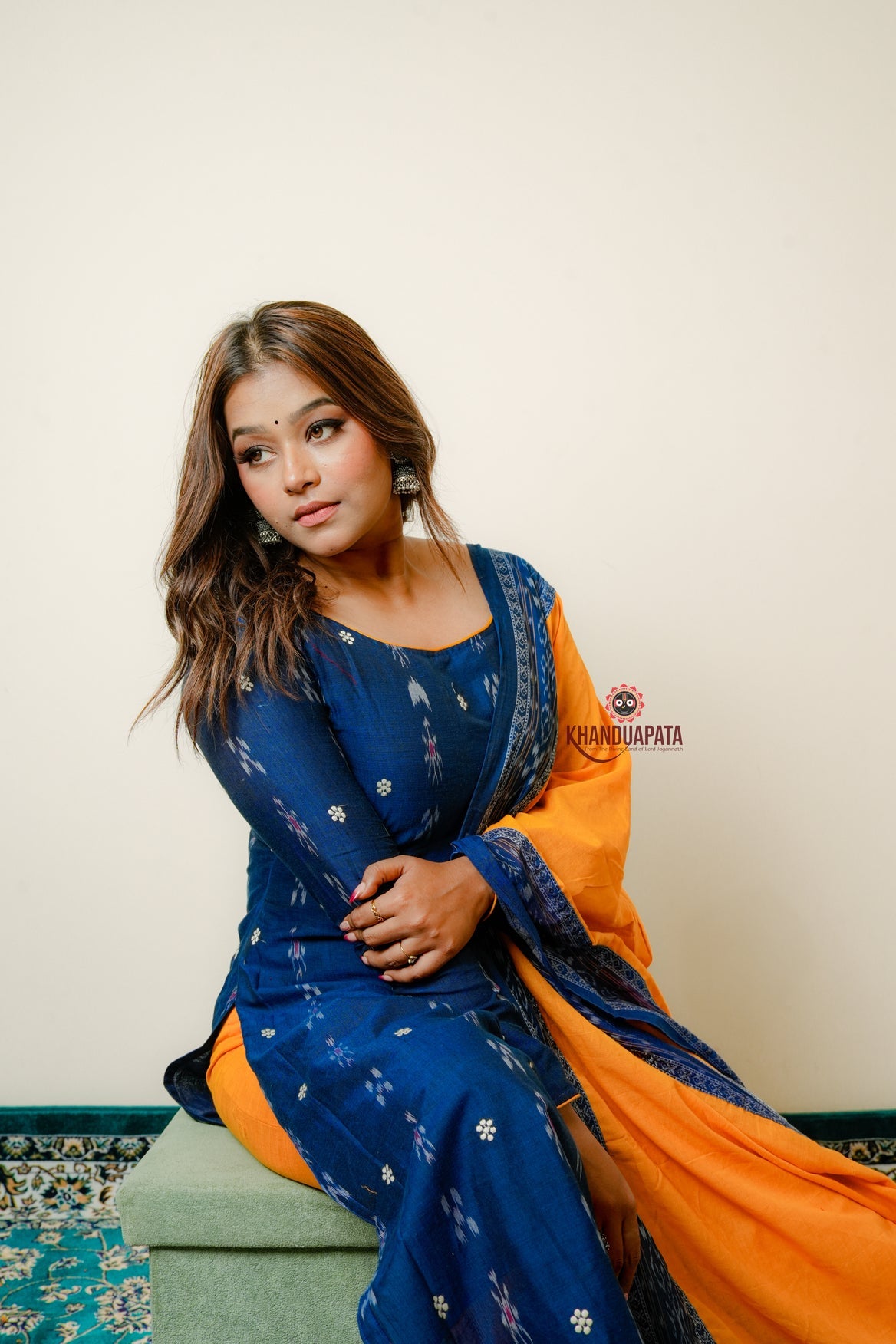Woman in a blue and orange traditional outfit sitting on a green cushion against a white wall.