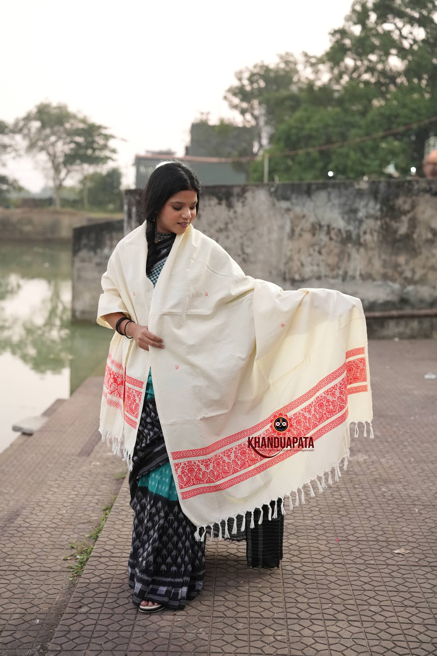 Woman holding a white shawl with red patterns by a body of water