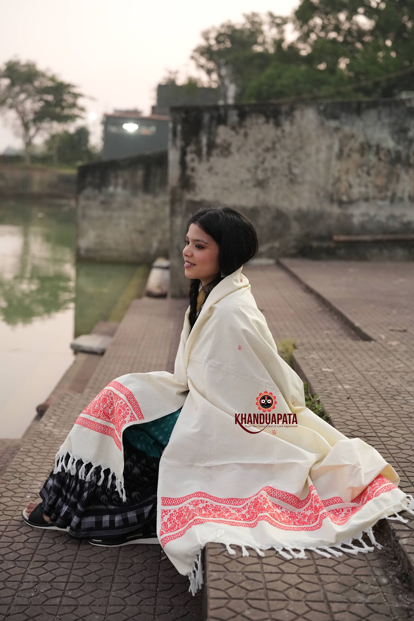 Woman sitting by a river wearing a white saree with red patterns, brand 'Khandudapata' visible.