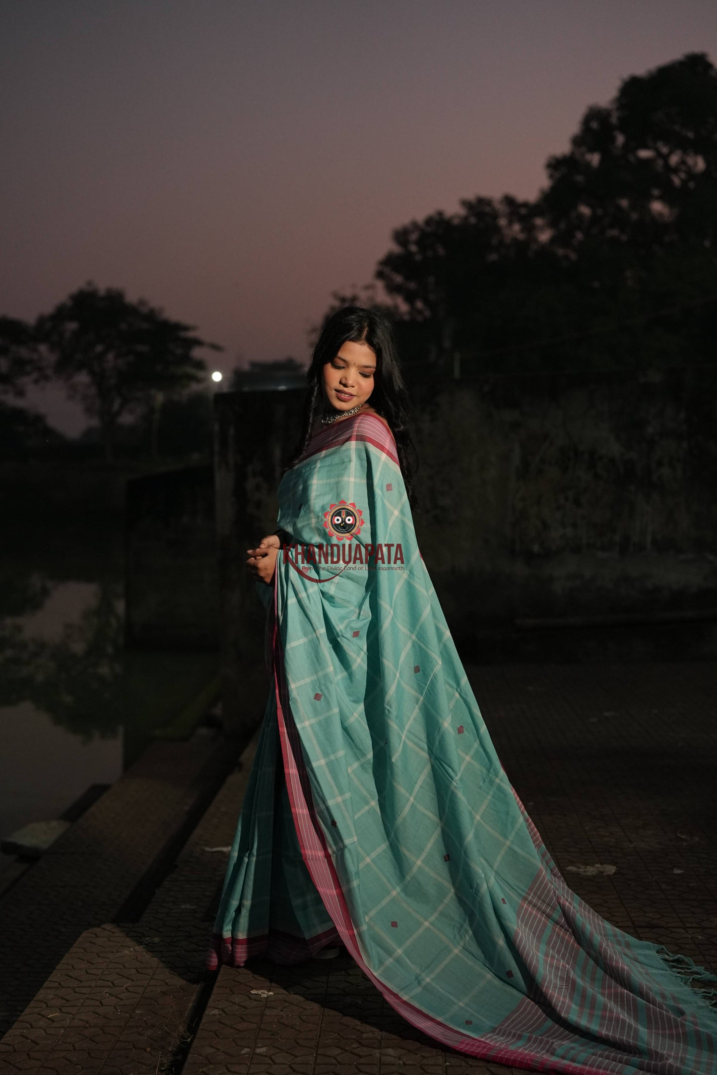Woman in a teal saree with pink border standing by a body of water at dusk.