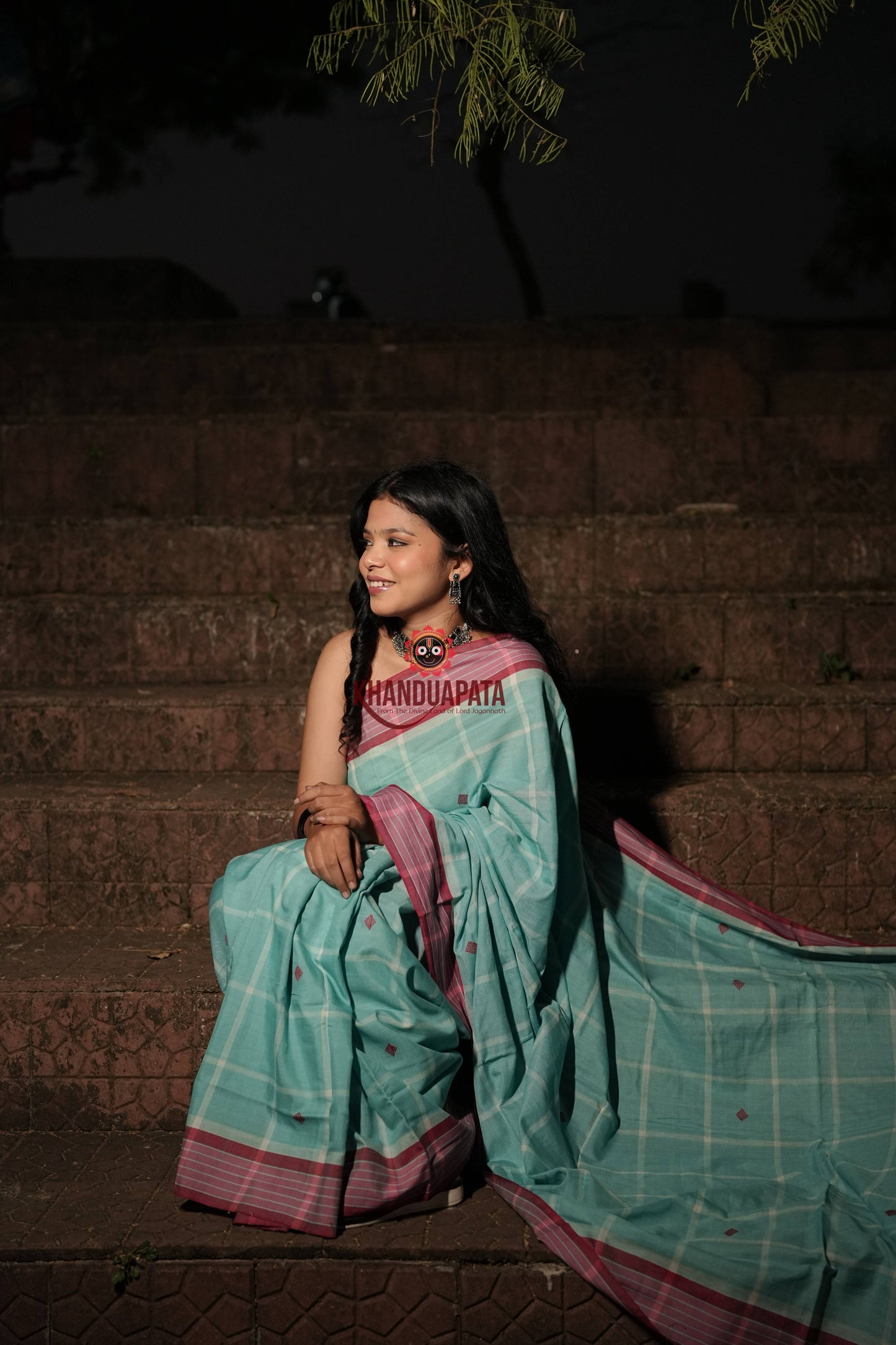 Woman in a teal saree with pink border sitting on steps at night.