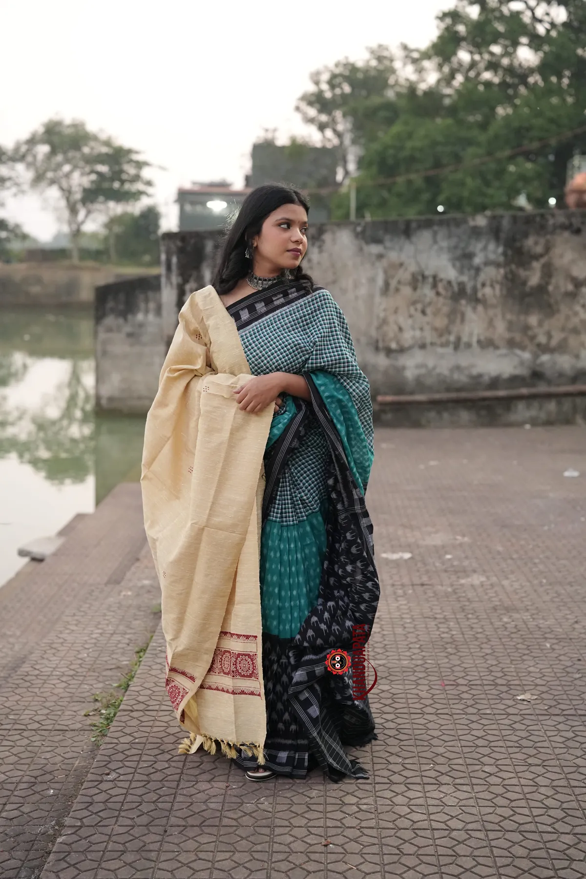 Woman in a traditional saree standing by a body of water with a stone wall and trees in the background.