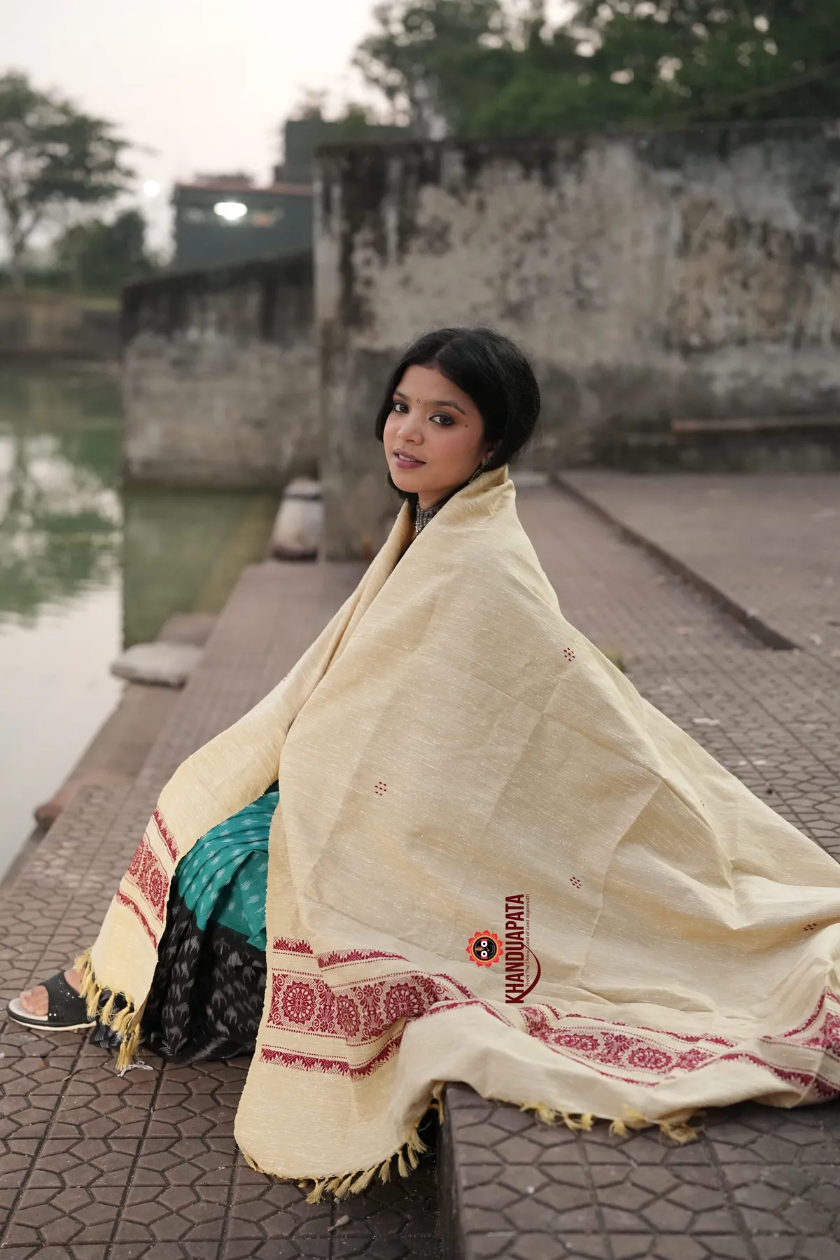 Woman sitting by a river with a cream-colored shawl draped over her, featuring a brand logo.