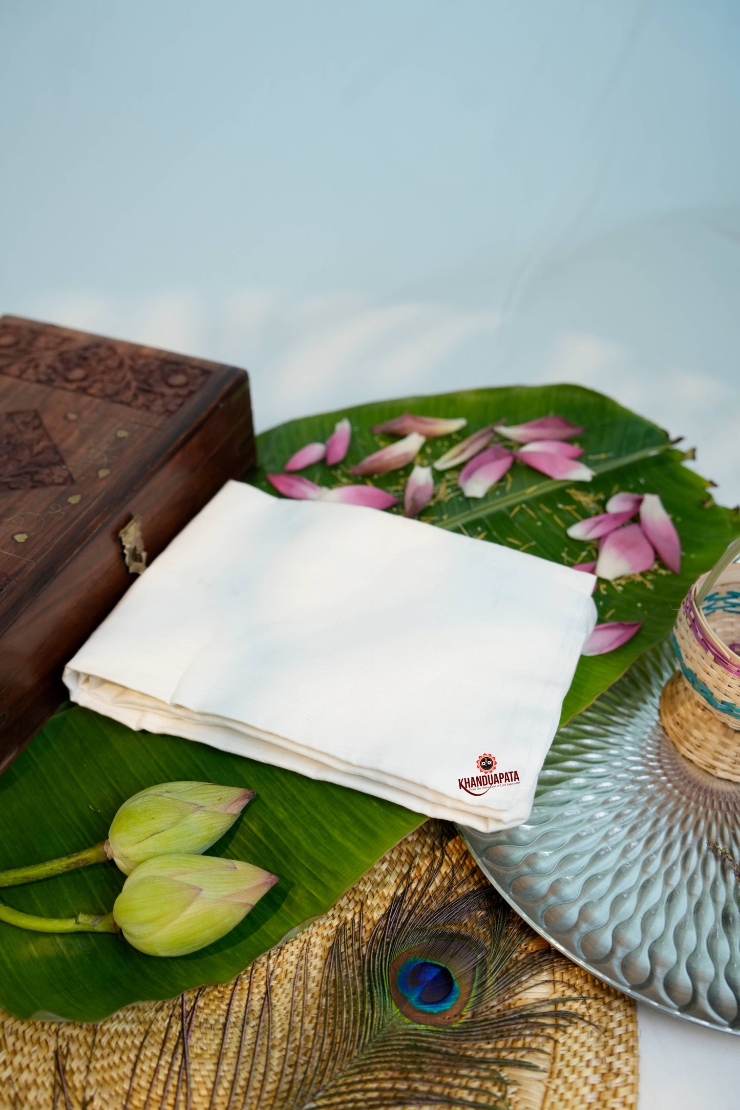 Wooden box, white cloth with logo, green leaves, pink petals, and peacock feather on a textured surface.