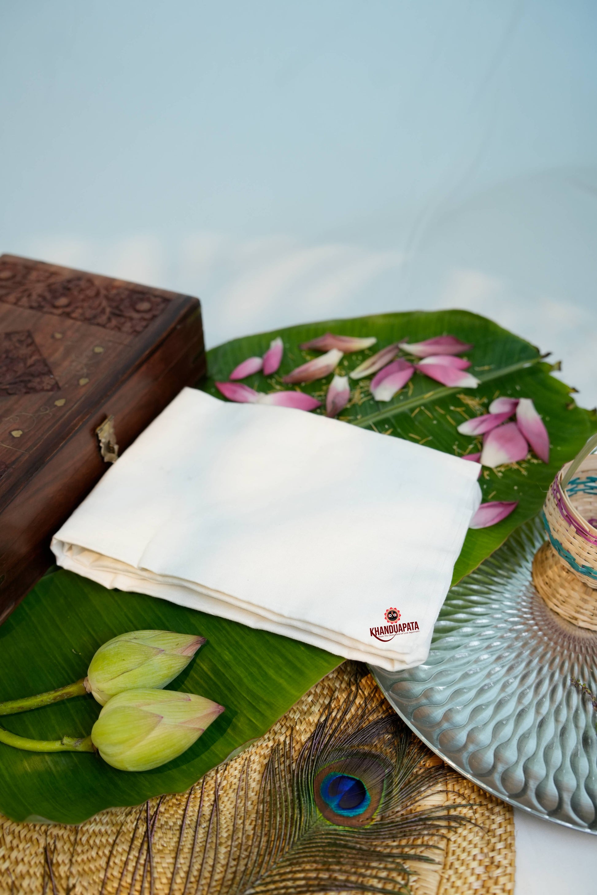 Wooden box, white cloth with logo, green leaves, pink petals, and peacock feather on a textured surface.